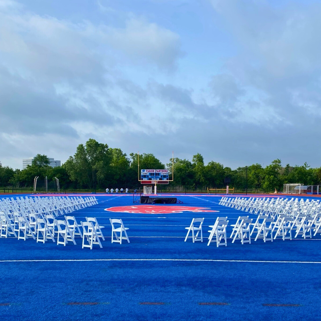 Outdoor graduation ceremony setup with stage and rows of white chairs on a field by TLC Event Rentals